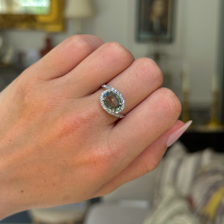 Green sapphire and diamond ring worn on left middle finger, closed hand in frame and bokeh antiquated background